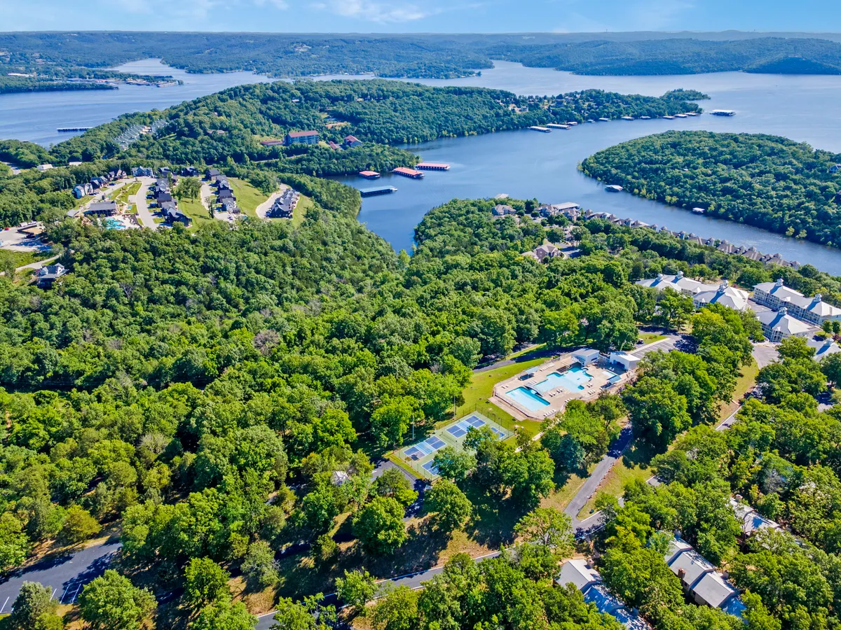 An elevated, panoramic view overlooking swimming pools, tennis courts, lush green landscaping, and Table Rock Lake with surrounding hills. The image highlights the resort’s proximity to the lake and its expansive recreational offerings.