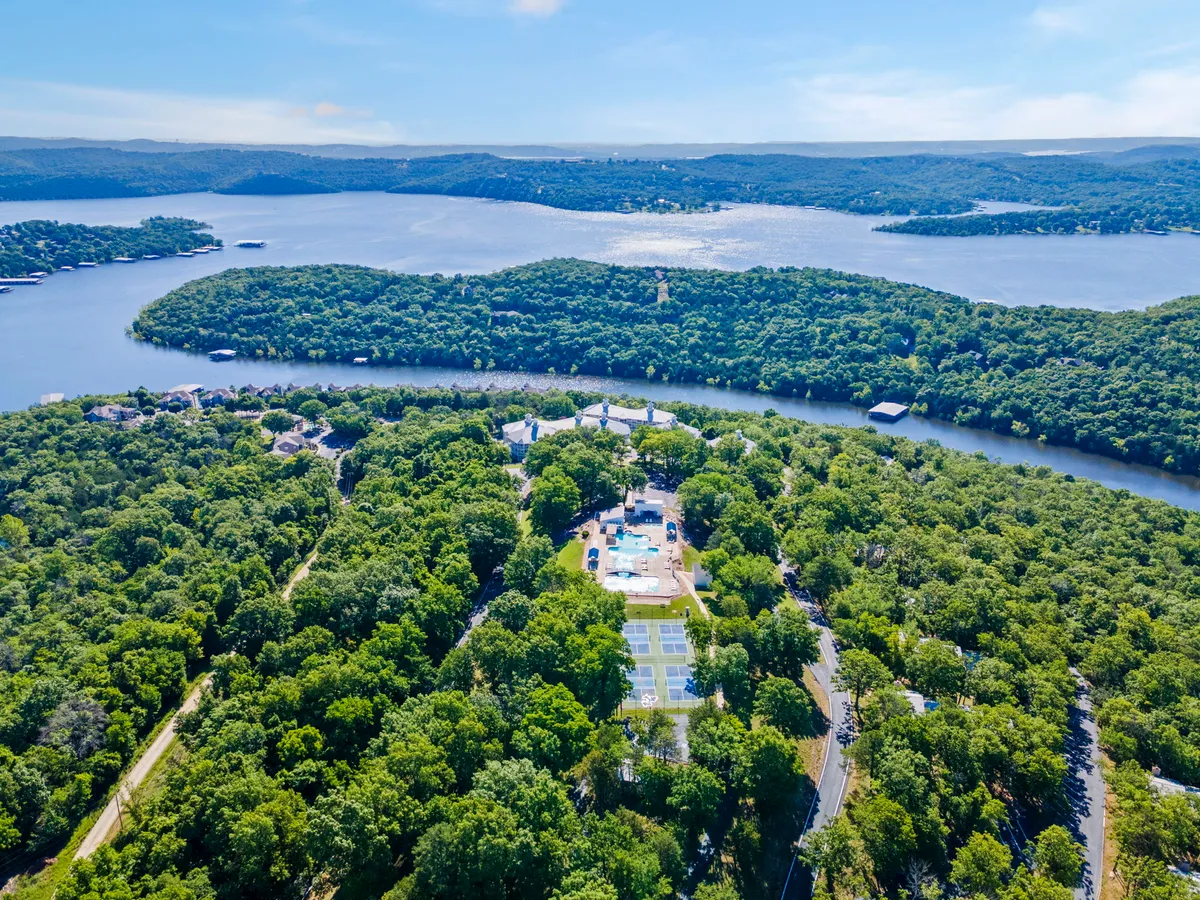 A panoramic view captures a wide expanse of water with lush greenery all around, a swimming pool area, and tennis courts in the distance. The scene is vibrant yet tranquil, showing both the natural beauty and recreational amenities available on the property.