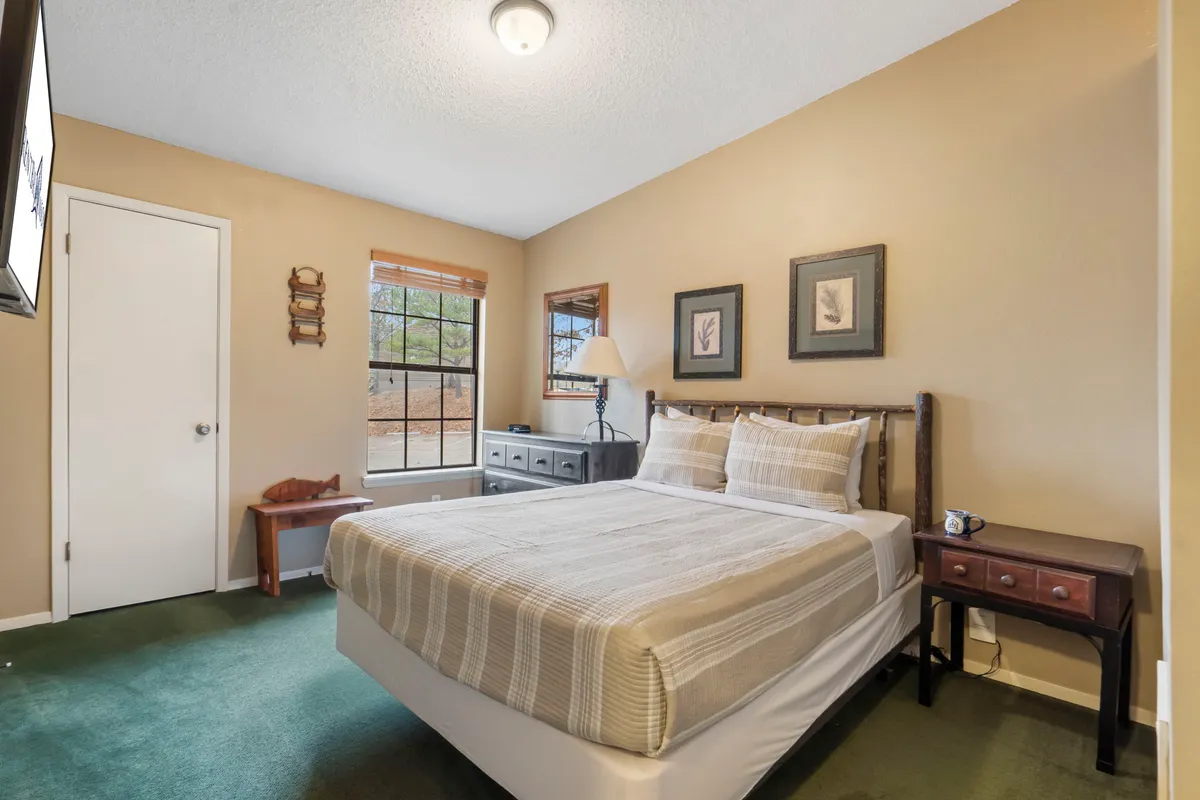 A calm and inviting bedroom is shown, featuring a neatly made bed framed by a large window. Soft natural light gently filters in, highlighting a dresser and a piece of framed art on the wall. The room feels peaceful and is designed for comfort and relaxation.