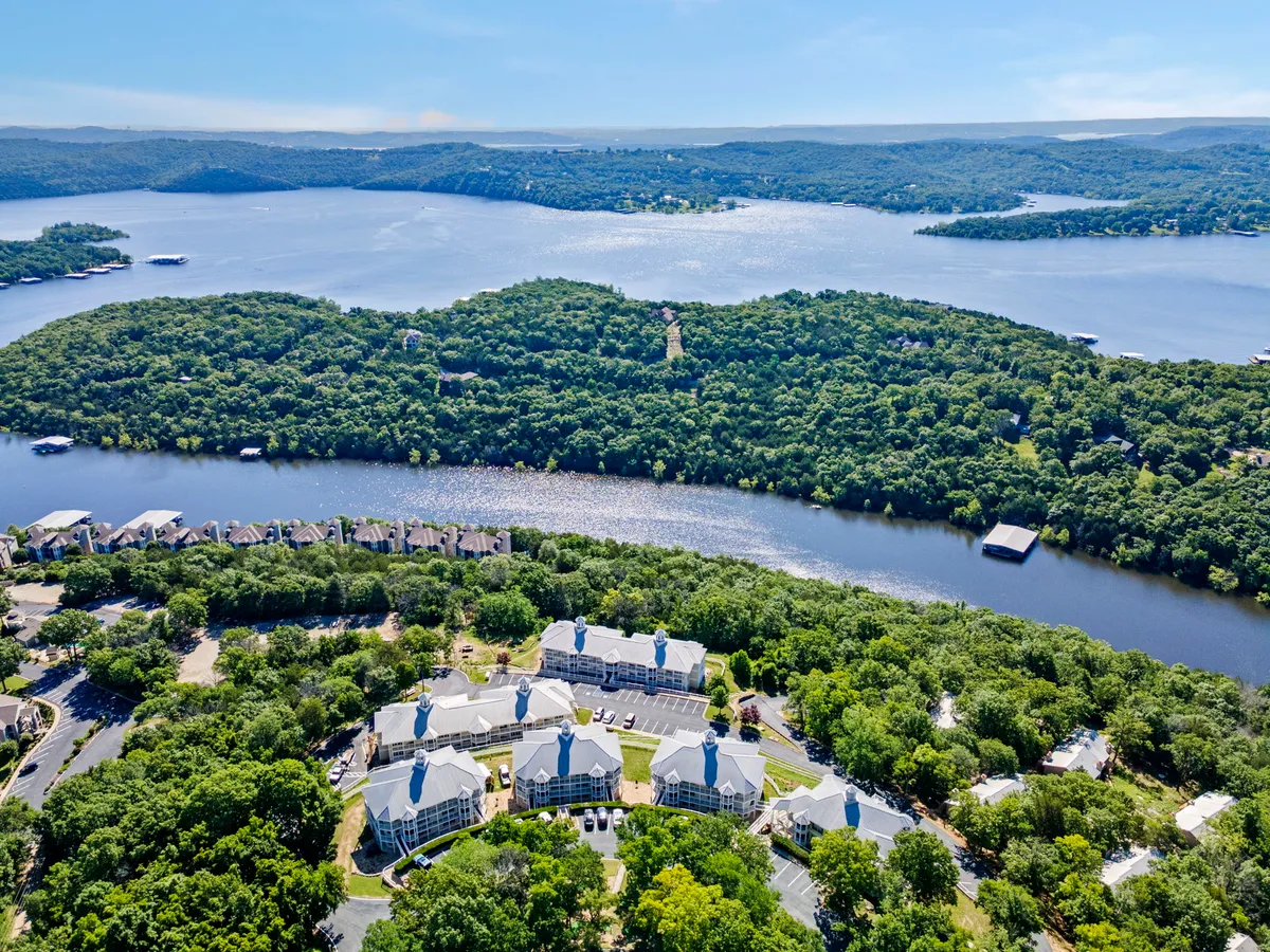 A tranquil exterior scene highlighting a peaceful lake set against wooded hills, with residential buildings nestled nearby. The lake is perfect for boating, fishing, kayaking, and other water activities, while the natural setting invites exploration year-round.
