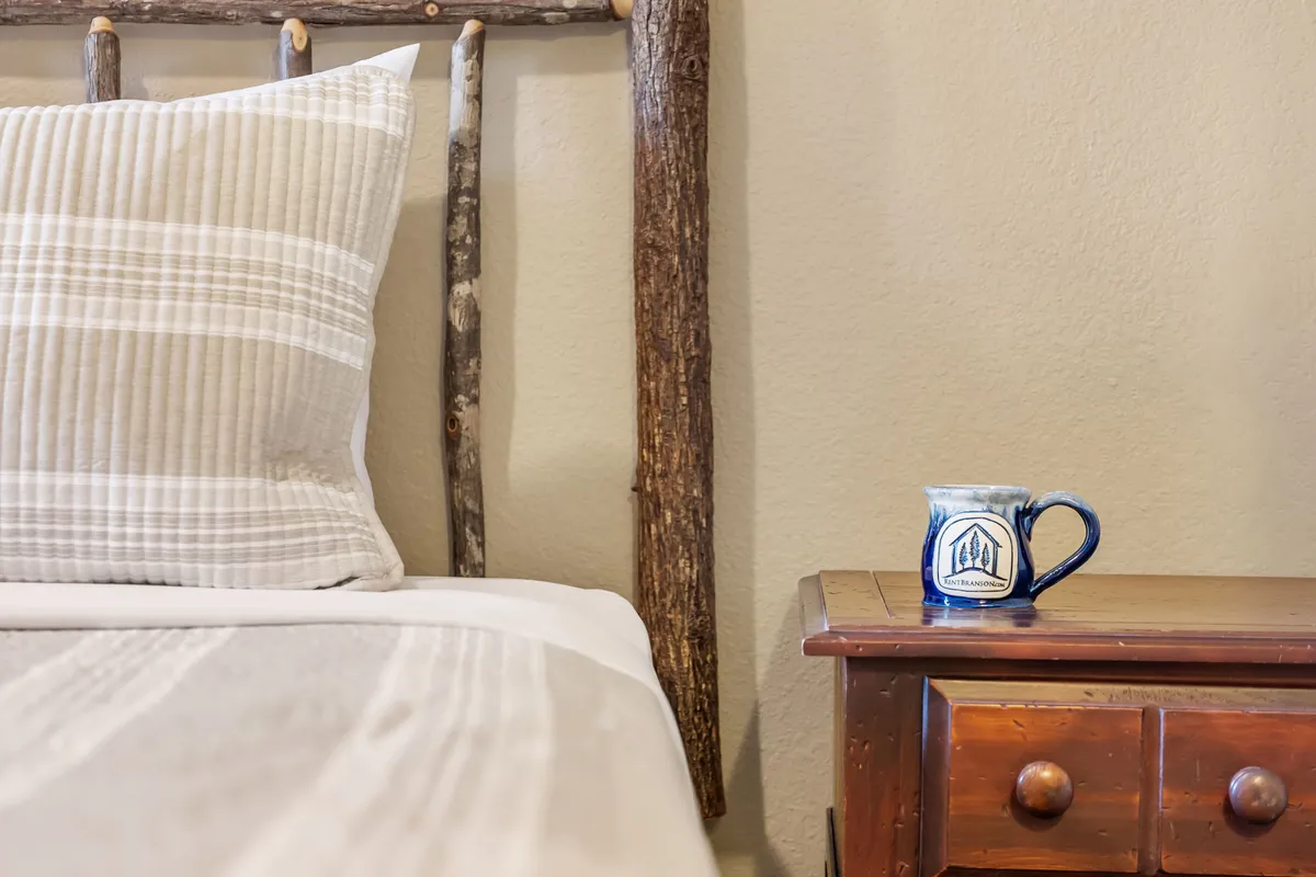 A log-framed bed with a striped pillow sits next to a wooden nightstand holding a ceramic mug. Gentle natural light streams through the window, illuminating the cozy space. The room exudes warmth and a welcoming, rustic charm.