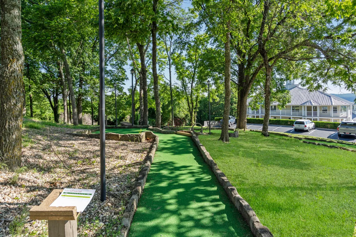 An exterior view of a mini-golf path winding through lush trees and marked by clear signage, evoking a playful and welcoming atmosphere. Families or groups can be imagined enjoying friendly competition as they navigate the course, with laughter and shared moments in focus.
