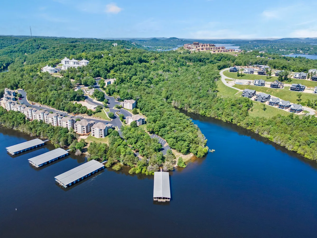 A serene view of the lakefront, featuring docks extending over the water, lush greenery, and residential buildings on a hillside. The environment feels peaceful, emphasizing easy lake access for boating and water activities.