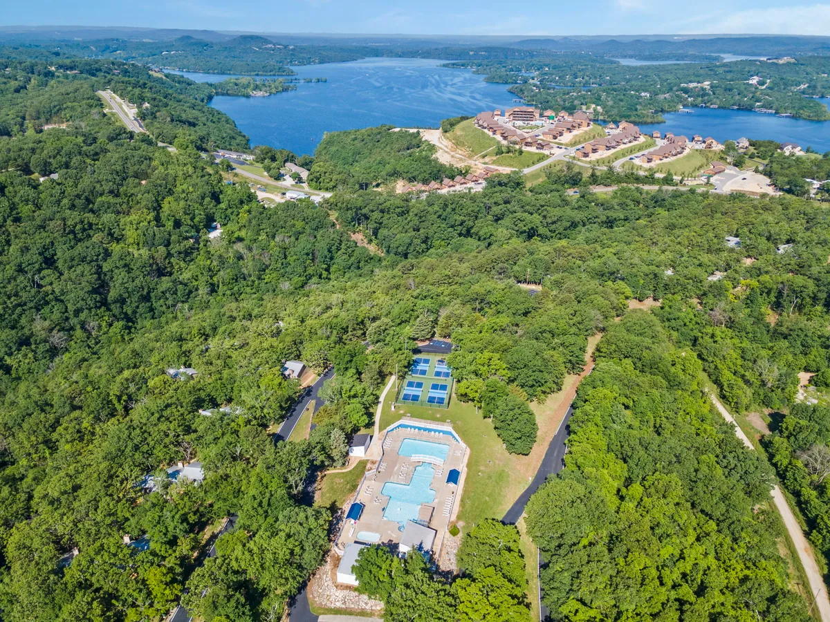 This exterior image shows a vibrant outdoor swimming pool area surrounded by lush green trees, with scenic waterfront views and resort buildings visible in the background. The inviting pool is the focal point, while the winding roads and open spaces suggest opportunities for exploration and outdoor activities.