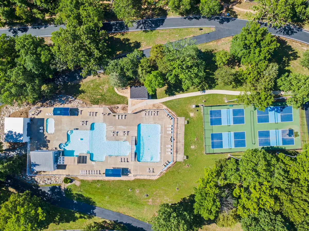 A vibrant exterior shot of a large swimming pool surrounded by numerous lounge chairs, with tennis courts visible in the background. The pool area is lively yet relaxing, inviting guests to enjoy swimming, sunbathing, or socializing. The scene is framed by mature trees, lending a sense of seclusion and resort ambiance.