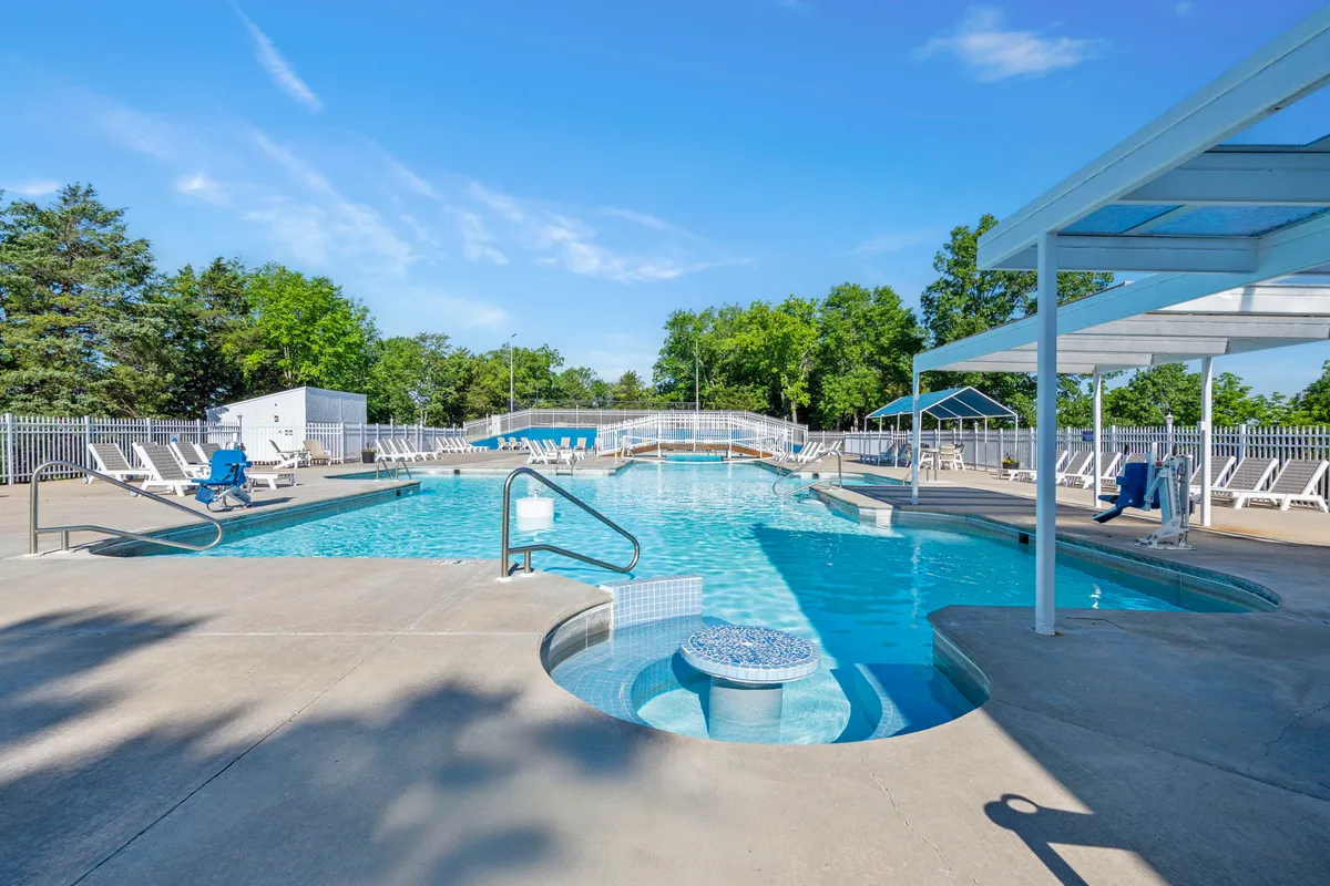A sparkling outdoor swimming pool with an adjacent hot tub, several lounge chairs, and families socializing and relaxing in the sun. The setting suggests a bustling yet relaxed community atmosphere perfect for summer days and poolside gatherings.