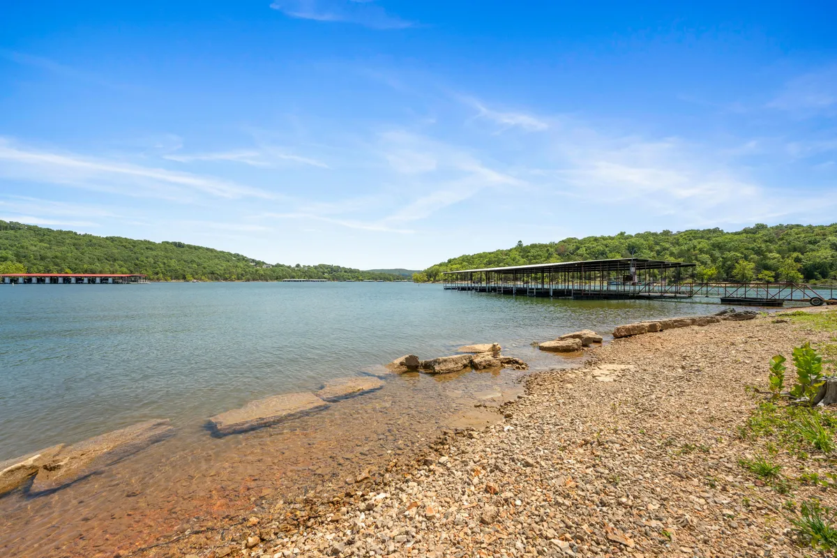 This image presents a tranquil lakeside setting with a dock extending into the water, a rocky shoreline, and lush surrounding trees. The calm lake is central, inviting thoughts of boating, fishing, and swimming, while the natural beauty of the Ozarks is captured in the background.