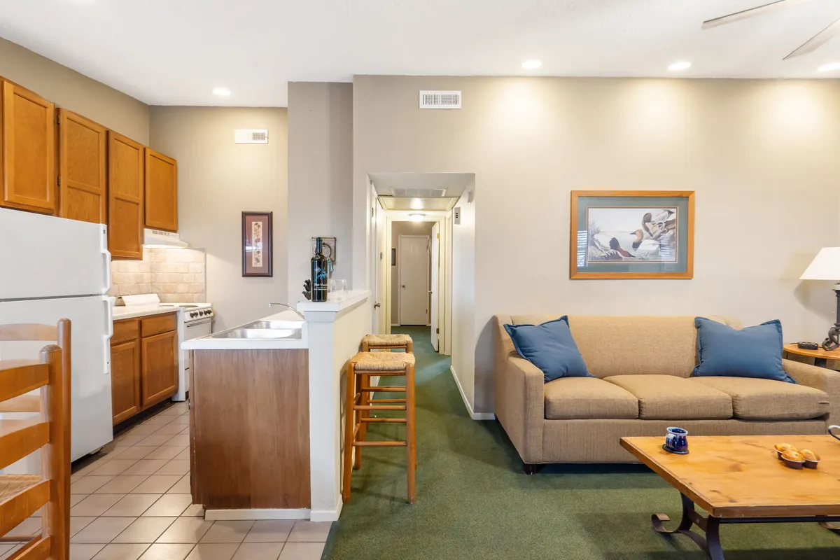 A cozy interior shot with a sofa in the foreground, a welcoming kitchen area with dining stools behind, and art on the wall. The arrangement suggests seamless interaction between cooking, dining, and lounging, reinforcing the open-plan living concept.